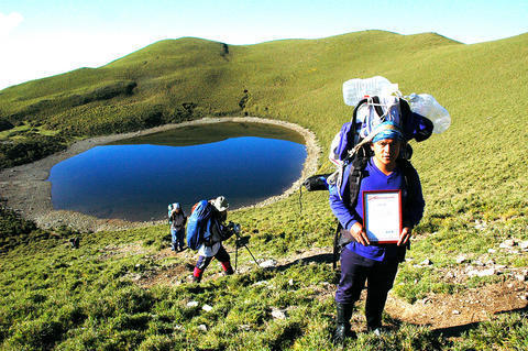 Chia-ming Lake (aka Meteor Lake) in Taiwan. Two hikers are in the foreground with a small deep blue  lake behind them. Green grass covers the crater around the lake.