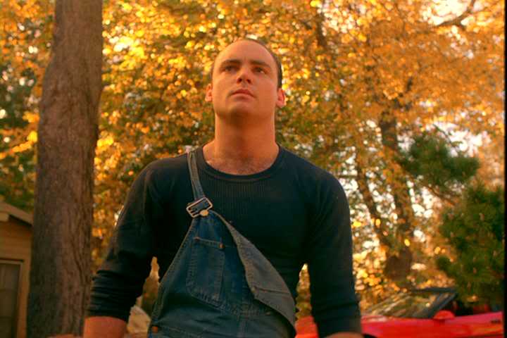 Leo Johnson from Twin Peaks. Leo stands in a street wearing coveralls, while the autumn leaves on the trees are  mostly yellow.