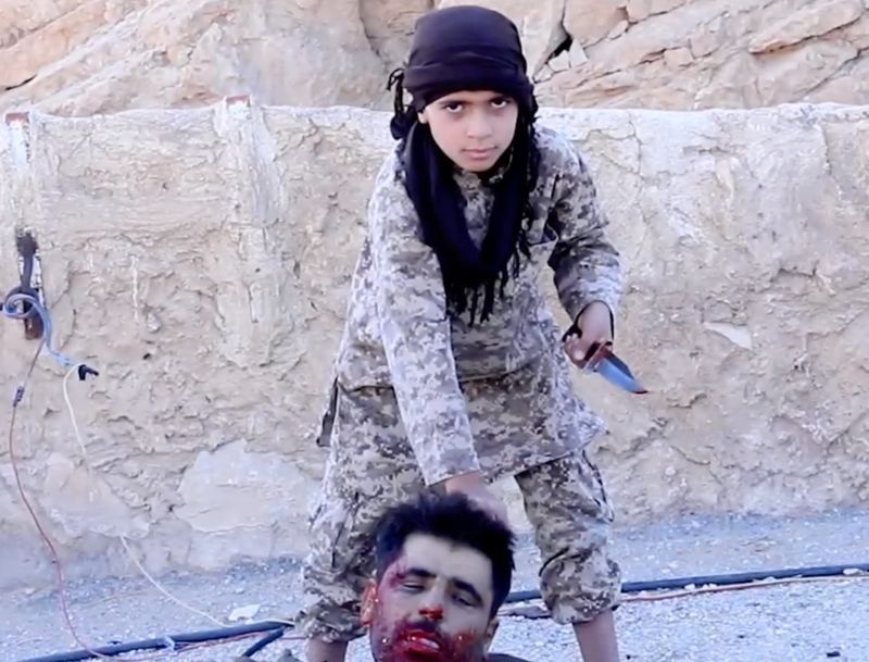 10 year old boy in ISIS territory holds dagger in his left hand and a severed human head in his right.