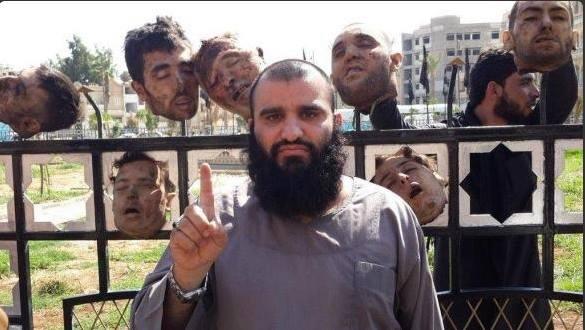 Fat ISIS terrorist raising 1 finger in front of iron fence with 5 human heads impaled on fence spikes. A man in the background adds 2 more heads to the grisly scene.